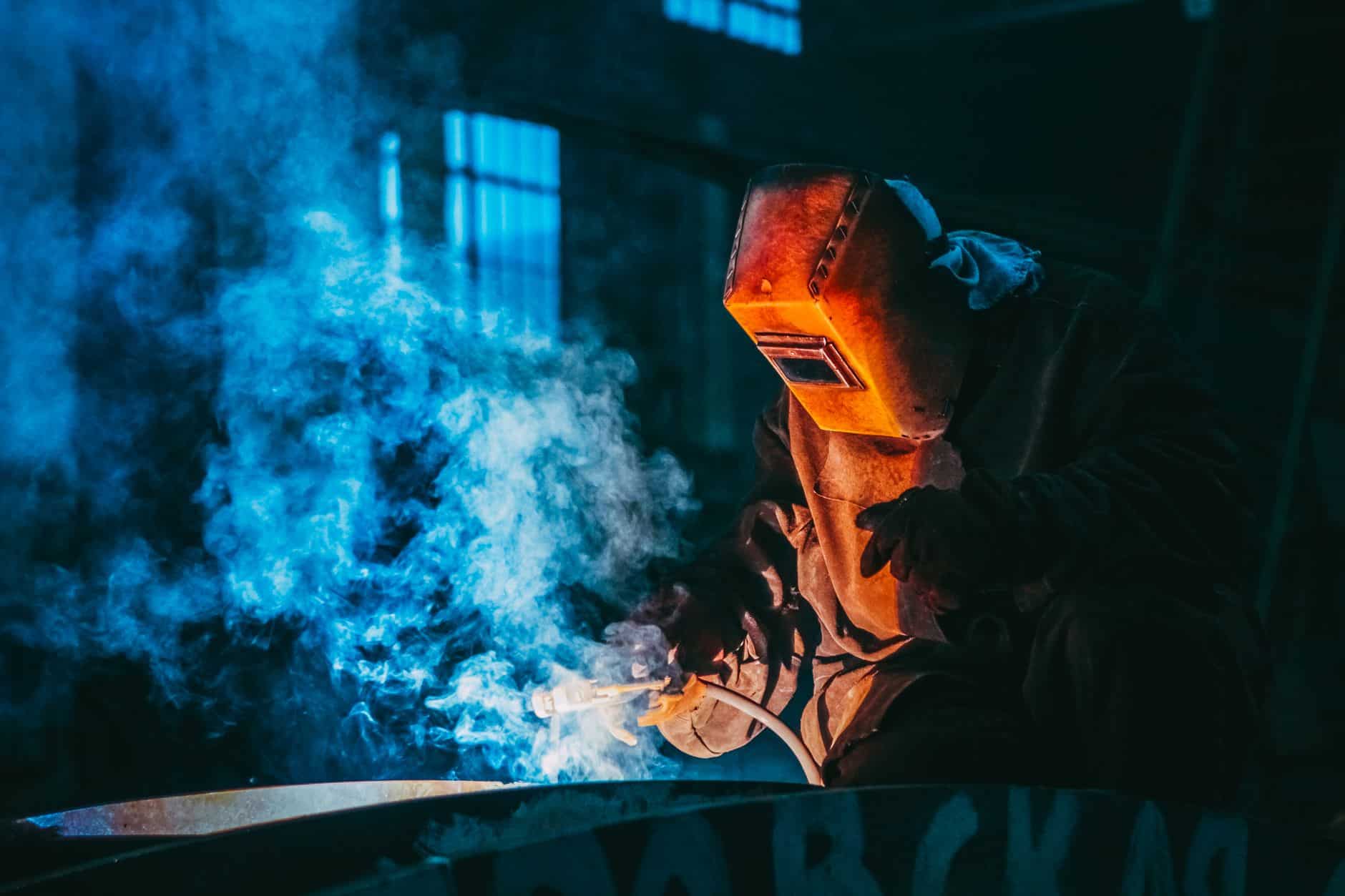 Welder in protective gear working amid blue welding smoke in a dark industrial workshop